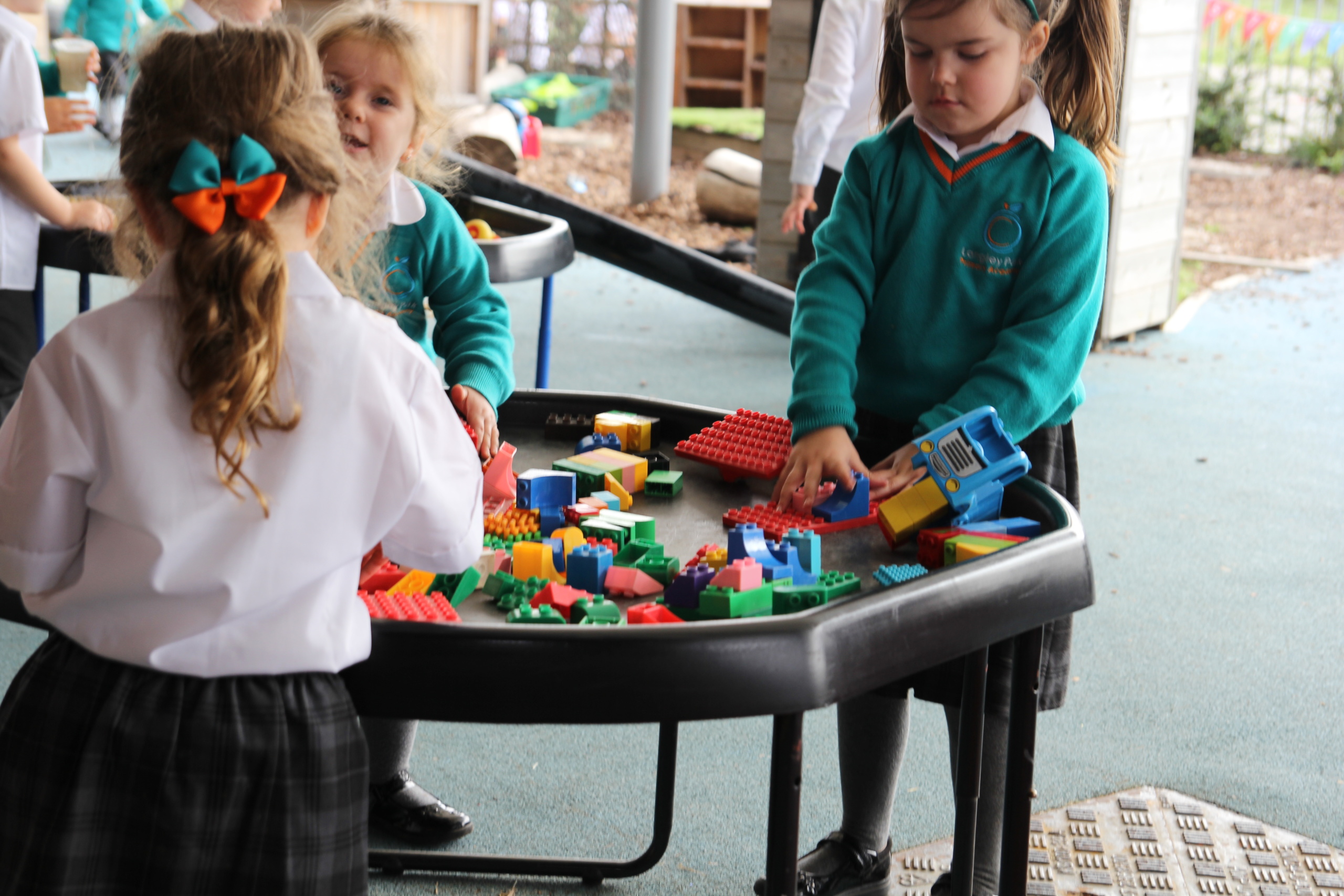Three young children stand around a table and play with colourful bricks outside