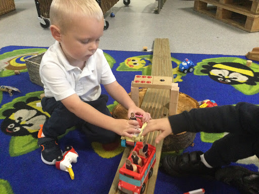 Two young male pupils are pictured sat together on a carpeted area of the classroom, playing with wooden blocks they have made into a bridge and some toy vehicles.