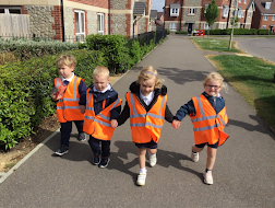 Four young pupils wearing orange-coloured fluorescent jackets are pictured walking hand-in-hand together down a pavement beside the road.